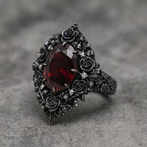 Puede incluir: Un anillo de plata oxidada con una gran piedra preciosa de color rojo intenso y corte ovalado en el centro. La banda y el engaste están intrincadamente esculpidos con motivos de rosas y pequeñas piedras claras incrustadas en el metal.
