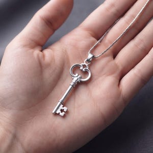 May include: A silver-toned metal key pendant suspended from a matching box chain necklace. The key features an ornate, rounded bow at the top, a slender cylindrical shaft, and a bit with a distinct cross-shaped cutout at the bottom end.