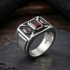 May include: A silver ring featuring a large rectangular deep red gemstone in the center, flanked by two smaller marquise-cut red stones. The metal band has a polished finish with recessed dark detailing around the stone settings and a smooth shank.