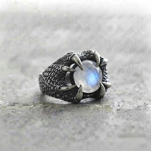 Könnte beinhalten: Ein silberfarbener Metallring mit einem strukturierten, schuppenartigen Band in Klauenform. Die Krallen halten einen runden, durchscheinenden Mondstein-Cabochon, der in der Mitte einen sanften, irisierenden blauen Schimmer aufweist.