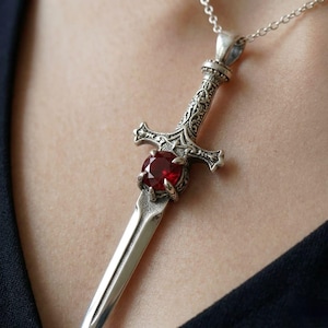 Può includere: Un ciondolo in metallo color argento a forma di spada, caratterizzato da un'impugnatura finemente lavorata e da una pietra preziosa rotonda sfaccettata di colore rosso intenso incastonata sulla guardia. Il ciondolo pende da una catenina.