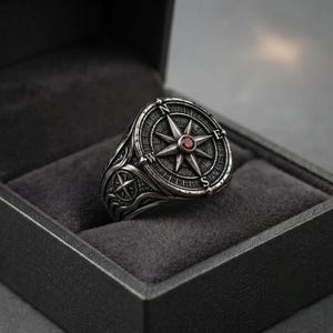 May include: A silver-toned metal ring featuring a circular compass rose design on the face. The center of the compass is set with a small, round, deep red gemstone. The band is engraved with directional letters N, E, S, and W and wave patterns.