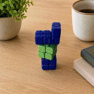 Peut inclure: Un jouet anti-stress composé de cubes interconnectés dans des tons bleu foncé et vert clair. Le jouet présente un motif texturé en forme de grille sur chaque face de cube et est disposé en une pile verticale légèrement décalée sur une surface en bois.