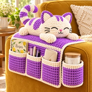Puede incluir: Un organizador de reposabrazos de ganchillo con una figura de gato a rayas crema y morado encima. El organizador tiene cuatro bolsillos en morado y crema, que contienen un libro de recetas, tres bolígrafos, una cartera y una vela en tarro.