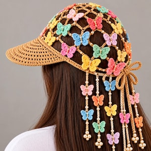 Può includere: Un berretto da baseball all'uncinetto color marrone chiaro con un motivo a reticolo decorato con numerose piccole farfalle multicolori nelle tonalità del rosa, blu, giallo, verde e arancione. Fili pendenti con farfalle e perline bianche pendono dal retro.