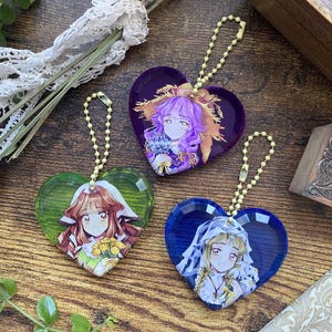Puede incluir: Tres dijes de acrílico en forma de corazón con bordes facetados y cadenas de bolas amarillas. Cada dije presenta una ilustración de personaje estilo anime diferente: uno con cabello morado y lavanda, uno con cabello castaño y flores amarillas, y uno rubio.