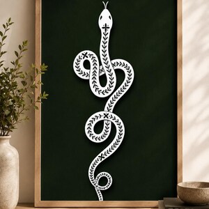 May include: A framed wall art piece featuring a white snake illustration on a dark green background. The snake is depicted in a vertical, coiled position and is decorated with a repeating black leaf pattern along its entire body length.