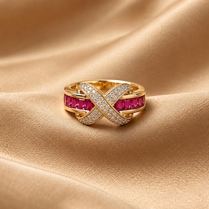 Könnte beinhalten: Ein goldener Ring mit einem unendlichkeitsförmigen Design, das mit kleinen, klaren, runden Steinen besetzt ist. Das Band enthält auf beiden Seiten Reihen von rechteckigen, leuchtend roten Steinen, die einen Kontrast zu den klaren Steinen bilden.