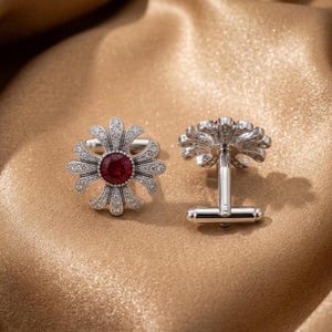 Puede incluir: Un par de gemelos plateados con un diseño floral que presenta una piedra preciosa roja redonda en el centro, rodeada de pequeñas piedras claras engastadas en pavé. Los pétalos metálicos tienen textura detallada y el cierre es de tipo basculante.