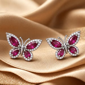 May include: A pair of butterfly-shaped stud earrings featuring vibrant marquise and pear-cut red stones set in silver-toned metal. Each wing is outlined with a border of small, clear, sparkling stones that catch the light, creating a detailed insect design.