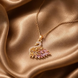 Può includere: Una collana color oro con un ciondolo a forma di cigno. Il collo del cigno è incastonato con piccole pietre chiare, mentre l'ala è composta da pietre taglio marquise in una sfumatura di tonalità gialle, arancioni e rosa su montatura dorata.