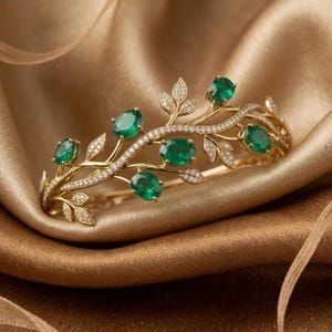 May include: A gold cuff bracelet featuring a vine-like design with five oval-cut green emerald gemstones and small pavé-set white diamonds along the branches and leaves, creating a detailed botanical structure with a polished metallic finish.