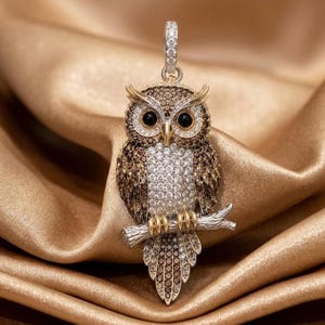 May include: An owl pendant featuring a detailed design with brown and clear pavé stones. The owl has black circular eyes, a gold-toned beak, and gold-toned talons gripping a silver-toned branch. A small bail at the top is encrusted with clear stones.