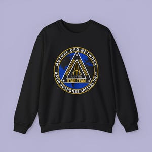 Puede incluir: Una sudadera negra de manga larga con un logotipo circular en el pecho. El logo muestra un fondo de globo terráqueo azul, un triángulo blanco con el texto INVESTIGATION, UFO RESEARCH y STAR TEAM, y el texto MUTUAL UFO NETWORK RAPID RESPONSE SPECIAL UNIT.