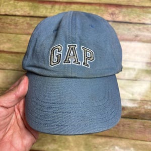 Peut inclure: Une casquette de baseball bleu clair avec le mot GAP brodé en blanc avec un contour foncé sur le panneau avant. La casquette présente une visière incurvée avec des coutures visibles et une calotte structurée en tissu de type coton résistant.