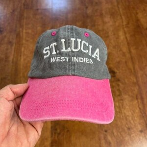 May include: A baseball cap with a washed grey crown and a bright pink brim. The front of the crown features white embroidered text that reads ST. LUCIA WEST INDIES. The cap includes pink eyelets on the top panels and decorative stitching on the brim.