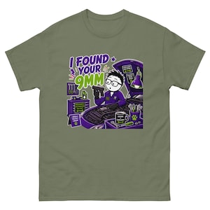 Puede incluir: Una camiseta verde oliva con un gráfico de dibujos animados de un mecánico trabajando en el motor de un coche morado. El texto dice "I FOUND YOUR 9MM". La escena incluye herramientas, una taza con "CAFFEINE & COMPLAINING" y un cartel que dice "I BREAK IT YOU FIX IT I REPEAT."