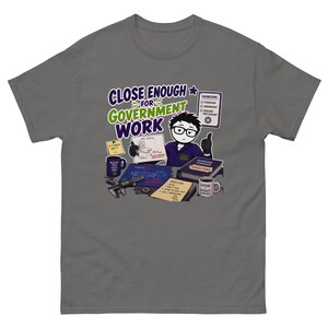 Puede incluir: Una camiseta gris carbón con una ilustración de dibujos animados de un ingeniero en un escritorio con el texto "CLOSE ENOUGH FOR GOVERNMENT WORK". El gráfico incluye planos, una taza con "CAFFEINE & INTEGRITY" y una lista "ENGINEERING".