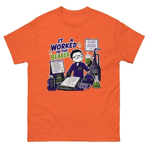 Puede incluir: Una camiseta naranja con una ilustración de dibujos animados de un químico con bata morada sosteniendo un vaso de precipitados verde. El gráfico incluye el texto "IT WORKED IN THE BEAKER", "CHEMICAL ENGINEERING", "TODAY'S PLAN" y "GOOD CHEMISTS FOLLOW RULES."
