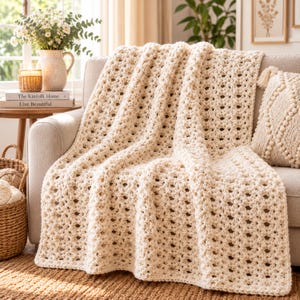 May include: A cream-colored, chunky crochet throw blanket with a repeating open-work stitch pattern. The blanket is draped over a sofa, showcasing its thick, textured yarn construction and uniform, rectangular grid design throughout the fabric.