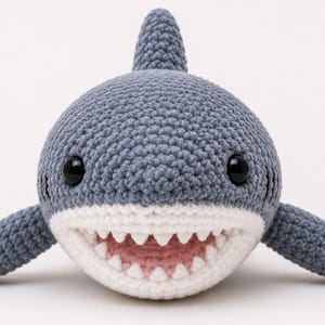 May include: A gray crocheted shark plush toy with a white underbelly and mouth area. The shark features two black plastic eyes, a dorsal fin on top, two side fins, and a wide open mouth showing a row of small white triangular teeth and a pink interior.