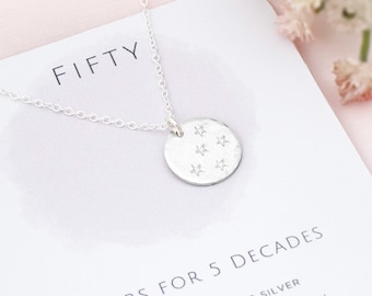 May include: A silver necklace with a round pendant featuring five stars. The pendant is hammered and has a textured surface. The necklace is on a white card with the text "FIFTY" and "5 STARS FOR 5 DECADES" in black lettering. The card also includes the text "MADE - STERLING SILVER" and "WISHEDFOR.CO.UK".
