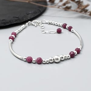 May include: A silver bracelet with a delicate design, featuring small silver beads and faceted, deep red gemstones. The bracelet has a curved bar and an adjustable chain clasp. The gemstones are evenly spaced along the bracelet.