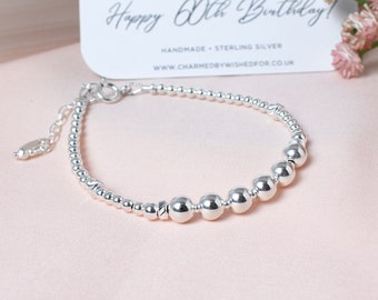 May include: A silver bracelet with small beads and larger beads. The larger beads are arranged in a line with a small decorative bead in the center. The bracelet has a lobster clasp closure. The bracelet is on a pink surface with a card that says "Happy 60th Birthday!" Handmade - Sterling Silver. www.charmedbywishedfor.co.uk