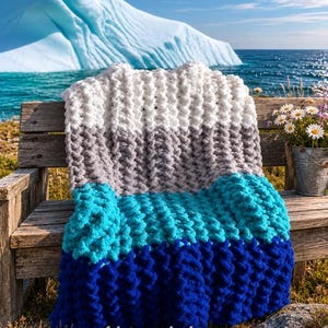 May include: A chunky knit blanket featuring horizontal color-blocked stripes in white, light gray, turquoise, and deep royal blue. The thick yarn creates a textured, looped pattern throughout the rectangular throw. The text "wild & wooly knits co" is visible at the bottom.