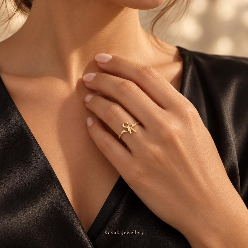 Puede incluir: Un anillo de oro que presenta las letras S y K en una fuente cursiva conectada y pulida. El anillo tiene una banda delgada y lisa, y se lleva en el dedo anular. El metal tiene un acabado c&aacute;lido y reflectante que captura la luz con brillo.