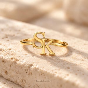 Puede incluir: Un anillo de tono dorado que presenta las letras S y K como punto focal central. La banda es delgada y pulida, con las letras may&uacute;sculas S y K conectadas para formar un elemento de dise&ntilde;o singular. El metal tiene un acabado liso y brillante.