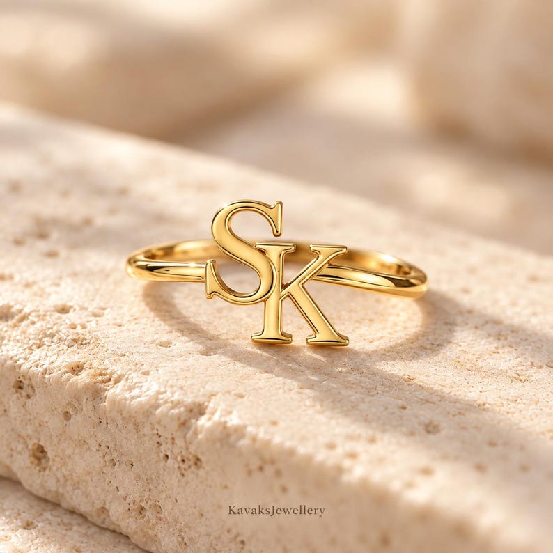 Puede incluir: Un anillo de tono dorado que presenta las letras S y K como punto focal central. La banda es delgada y pulida, con las letras may&uacute;sculas S y K conectadas para formar un elemento de dise&ntilde;o singular. El metal tiene un acabado liso y brillante.