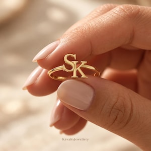 May include: A gold ring featuring the letters S and K as a central focal point. The letters are connected in a serif font style, forming a monogram design. The ring band is thin and polished, showcasing a smooth, reflective metallic finish throughout.