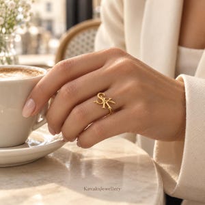 Puede incluir: Un anillo de oro con las letras S y K en una fuente estilizada. El anillo se lleva en un dedo, colocado junto a una taza de caf&eacute; y un plato de cer&aacute;mica blanca. Las letras est&aacute;n conectadas en una sola pieza con un acabado met&aacute;lico pulido.