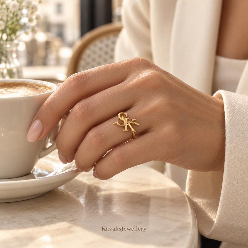 Puede incluir: Un anillo de oro con las letras S y K en una fuente estilizada. El anillo se lleva en un dedo, colocado junto a una taza de caf&eacute; y un plato de cer&aacute;mica blanca. Las letras est&aacute;n conectadas en una sola pieza con un acabado met&aacute;lico pulido.