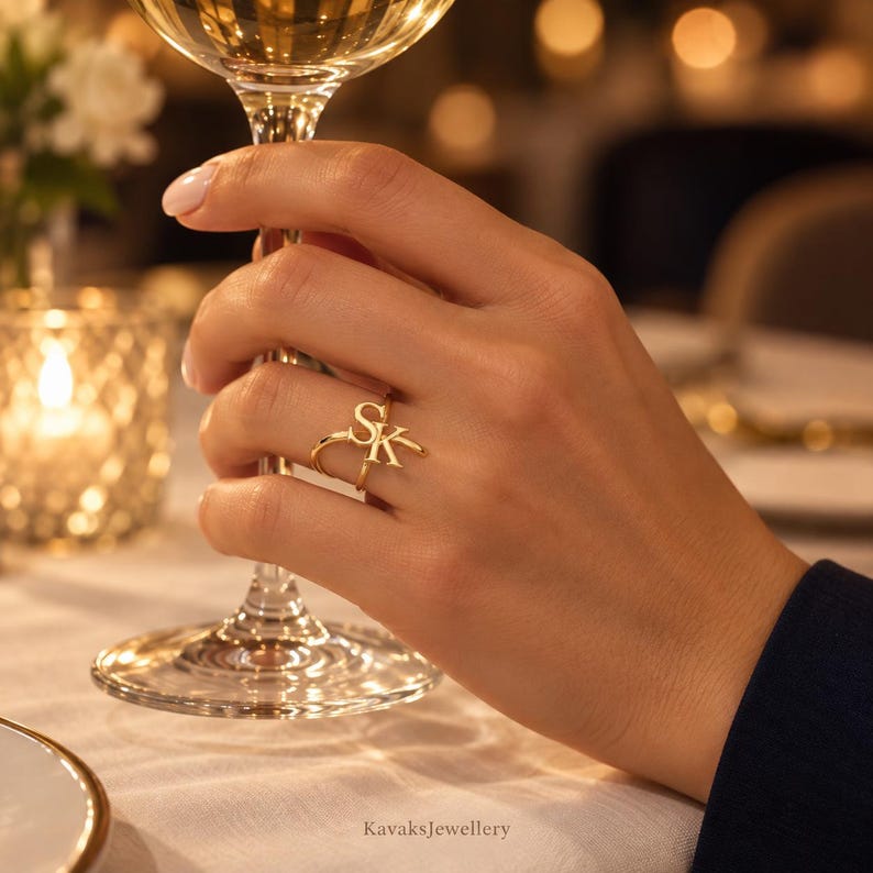 Puede incluir: Un anillo de tono dorado que presenta las letras S y K en una fuente cursiva estilizada. El anillo tiene una banda delgada y pulida que se envuelve alrededor del dedo, mostrando las letras may&uacute;sculas S y K entrelazadas como elemento central.