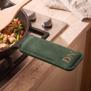 May include: A dark green leather skillet handle cover with the embossed letters DN, attached to a black cast iron pan filled with a mixture of cooked chicken, rice, green peas, and carrots, being stirred with a light-colored wooden spatula.