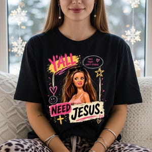 May include: A black t-shirt featuring a graphic of a doll with long brown hair and a pink top. The text reads Y'ALL NEED JESUS, PRAY. TRUST. DON'T STRESS. The design includes yellow and pink accents, a yellow star, a yellow cross, and hearts.