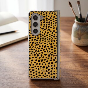 May include: A foldable smartphone case featuring a vibrant yellow background with a repeating black leopard print pattern. The case includes a vertical camera cutout on the left side and the word Arual printed in small black letters near the bottom.