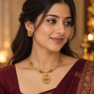 Può includere: Una collana color oro con un ciondolo circolare centrale raffigurante una divinità in rilievo, arricchita da piccole perline rosse e verdi lungo la catena, abbinata a orecchini a lobo circolari con un design in rilievo simile.