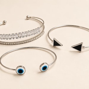 May include: Three silver-tone metal cuff bracelets. One features a double band with baguette and round clear crystals. Another is an open cuff with black triangular ends. The third is an open cuff with two circular evil eye charms.