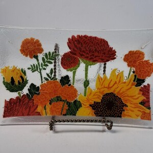 May include: A rectangular clear glass tray featuring a vibrant floral design with yellow sunflowers, orange marigolds, and deep red chrysanthemums. The glass has a textured surface with visible air bubbles and rests on a twisted metal stand.