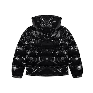 May include: A black puffer jacket with a glossy, patent-like finish, featuring a hood and horizontal quilted panels. The back of the jacket displays embroidered text in a gothic-style font across the upper section, with long sleeves and elastic cuffs.