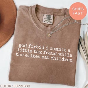 May include: An espresso-coloured short-sleeve t-shirt with a crew neckline. The shirt features white text printed on the chest that reads: god forbid i commit a little tax fraud while the elites eat children. A silver-toned necklace rests on the collar.