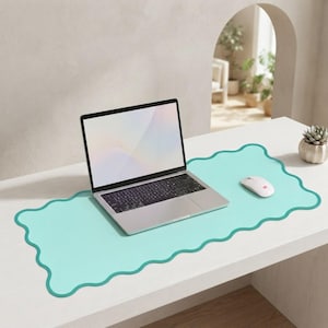 May include: A light mint green desk mat with a wavy, scalloped edge design. A silver laptop with a black keyboard and a white computer mouse are placed on top of the mat. The mat provides a smooth surface for the laptop and mouse to rest upon.