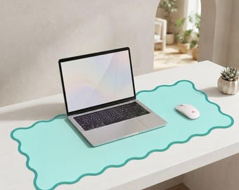 Peut inclure: Un tapis de bureau vert menthe clair avec un design à bords ondulés et festonnés. Un ordinateur portable argenté avec un clavier noir et une souris d'ordinateur blanche sont posés sur le tapis, offrant une surface lisse pour le matériel.