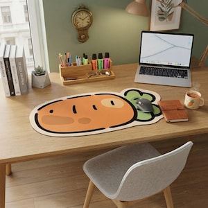 May include: A large, oval-shaped desk mat featuring a cartoon carrot graphic with a bright orange body and green leafy top. The mat has a thick black outline and is positioned on a wooden desk surface alongside a laptop, a notebook, and a mug.