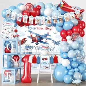 May include: A first birthday party decoration set featuring a balloon arch in red, white, and light blue, a backdrop with an airplane illustration and the text How time flies, a red number one balloon, and three light blue boxes spelling ONE.
