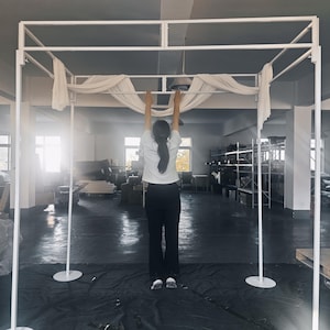 May include: A large, white, rectangular metal frame structure designed for event decoration. The frame features four vertical poles with circular base plates and a top canopy section draped with white fabric, creating a tall, open-air display.
