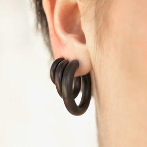 Könnte beinhalten: Dunkelbraune Holz-Creolen. Die Ohrringe sind kreisförmig und haben eine glatte, polierte Oberfläche. Die Ohrringe werden am Ohr getragen und sind dunkelbraun. Die Ohrringe sind ein schlichtes, aber elegantes Accessoire.